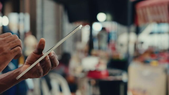 Hands of African American man using smart tablet at outdoor food market. Male service representative checks stock levels through app on handheld device. Guy manager reviews online feedback