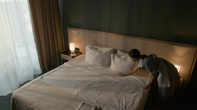 High angle view of hotel housekeeper arranging white sheets and pillows on bed in elegant guest room with bedside lamps and folded towels placed on blanket