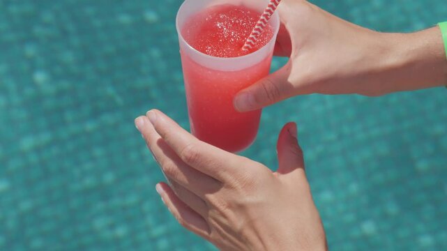 Woman's Hand with green wristband holding Plastic cup with pink cocktail drink and striped straw on turquoise water swimming pool on background. Relaxing in tropical hotel, Luxury holiday concept