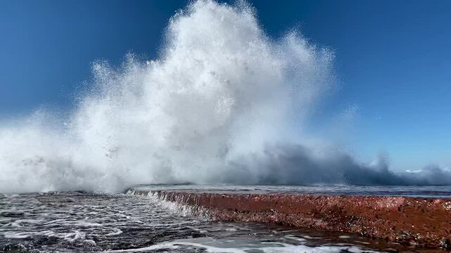 A giant wave hits an ocean rock wall with extreme force before exploding into a blue sky towards the camera. Captured in slow motion from in front of the the water explosion.