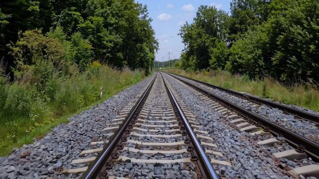 Low-angle video shot of parallel train tracks stretching into the distance, surrounded by lush greenery under a clear blue sky.