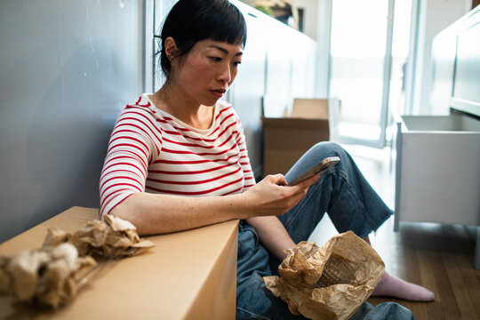 Woman sitting on floor checking phone while unpacking moving boxes at home