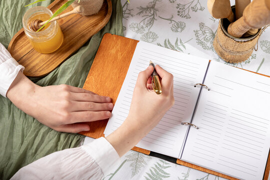 A woman writes a recipe in a wooden-cover notebook on a floral tablecloth, accompanied by a honey jar and pen. A cozy and inspiring workspace for creativity, journaling, and culinary planning.