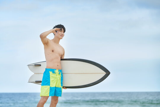 Young Asian Man on Beach Shielding Eyes While Holding Surfboard