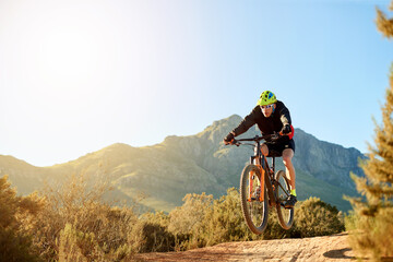 Fototapeta premium Man, mountain bike and jump with sunshine on dirt road for adrenaline rush, fitness or extreme sport in nature. Male person, cyclist or riding with bicycle for stunt or cardio on path, trail or space