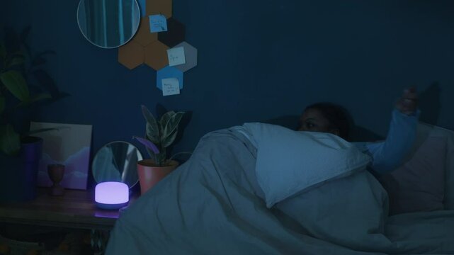 Young Black woman in pajamas getting out of bed and sleepwalking in dark bedroom at night