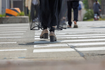 Low-angle person wearing light beige shoes and dark, sheer, black skirt-like garment walking across crosswalk marked by white painted lines on gray concrete. Angled perspective focuses on movement.