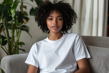 Young woman with curly hair wearing a plain white t-shirt sits comfortably on a gray sofa in a cozy living room surrounded by indoor plants, exuding a relaxed and inviting atmosphere