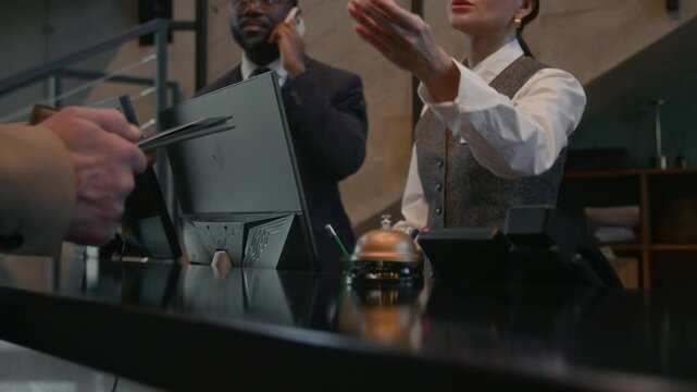 Female receptionist handing passport and credit card to guest after check in at hotel front desk while concierge speaking on phone in background