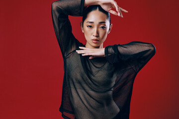 Fashion asian woman in sheer black top posing on red background with hands on hips and head tilted...