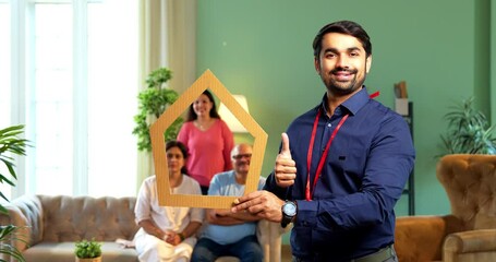 Holding house cutout and pointing, young Indian Asian male finance executive promotes real estate concept while happy family sits in background on sofa, symbolizing home loan, savings, and investment