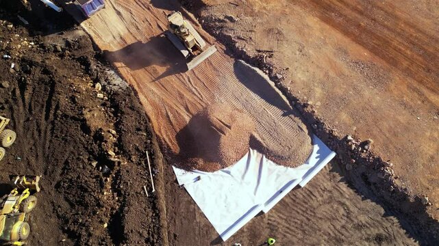 High angle top down late afternoon footage of bulldozer maching covering layers of geotextile with dirt and soil for the construction of a highway. Freeway build site with geosynthetic fabric burried
