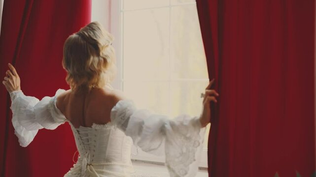 young woman queen opens curtains, uncovers the window looking out enjoy new day sun light. waiting for love. Fantasy fairy lady Happy girl blonde hair, white vintage dress old style back rear view 4k