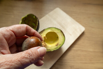 Hand holding avocado pit next to halved fruit on wooden cutting board.