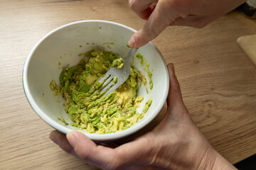 Mashing avocado with fork for guacamole in white bowl.