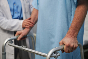 Doctor supporting senior patient using a medical walker in hospital, showing care and process of medical aid. Hands on walker, conveying professional assistance during recovery