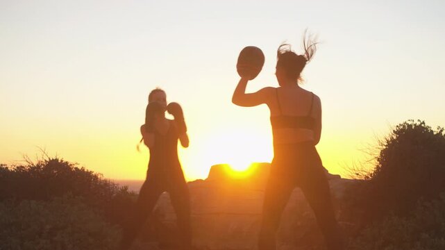 Sporty female fighter practicing boxing on hill overlooking seascape during sunset, with female coach guiding her. Determined boxer training in scenic beach setting