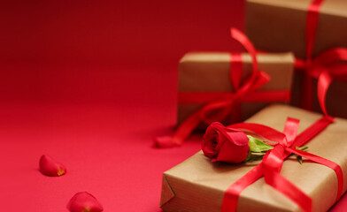 Elegant gift box wrapped in kraft paper with a red satin ribbon and decorated with a red rose on a vibrant red background. Romantic composition with soft focus, perfect for love-themed celebrations.