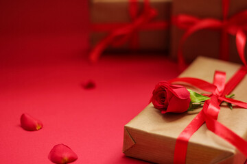 Elegant gift box wrapped in kraft paper with a red satin ribbon and decorated with a red rose on a vibrant red background. Romantic composition with soft focus, perfect for love-themed celebrations.