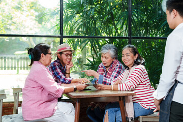 Senior Asian women group sitting casual cafe environment smiling waiter serving discussing ordering food happy conversation natural light greenery background enjoying retirement leisure lifestyle trip