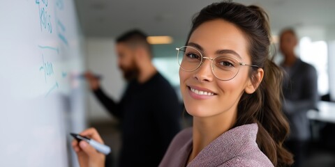 A woman wearing glasses is smiling at a white board with writing on it. The woman is the main focus of the image, and the white board is behind her
