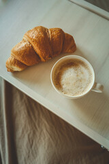 Morning breakfast in bed with fresh croissant and cappuccino on a white tray. Cozy neutral bedding and soft lighting create a relaxing start to the day