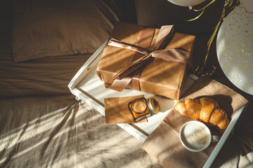 Cozy breakfast-in-bed scene with wrapped gift, candle, croissant, and coffee on a white tray, surrounded by warm sunlight and festive balloons, creating a soft, celebratory atmosphere
