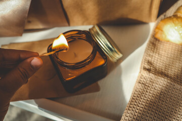Hand holding a lit match about to light a candle in a glass jar, next to a beautifully wrapped gift with a satin ribbon, captured in warm, golden morning light