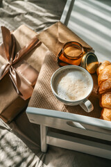 Breakfast in bed with a wrapped gift box, croissant, candle, and cup of coffee on a wooden tray, surrounded by soft bedding and festive balloons in beige and gold tones