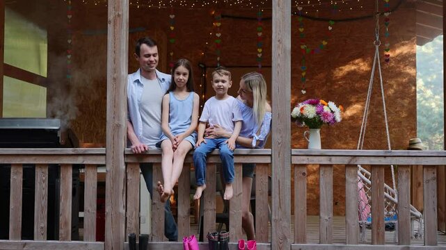 Loving family on veranda of house smiling happily. Portrait of mom, dad and boy, girl in country house, near grill being prepared. Concept of support, dreams, rental housing, own housing.