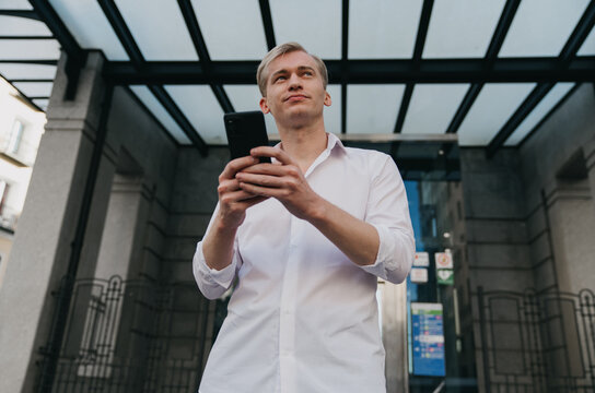 Focused young Caucasian businessman browsing smartphone outside modern office entrance, casual outfit, reflecting concentration, digital freelance work, career mobility, and tech innovation