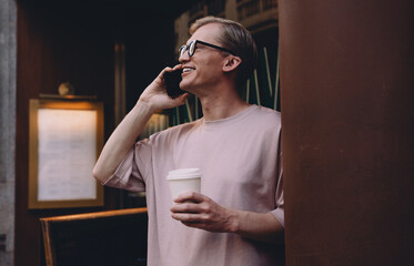 Young freelancer smiling while talking on smartphone, holding takeaway coffee, enjoying casual work outside near café with warm urban atmosphere and neon reflections in background