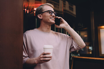 Stylish freelancer enjoying outdoor phone call, holding coffee cup, smiling and standing against urban neon reflections, representing mobile work freedom, casual career flexibility and positivity