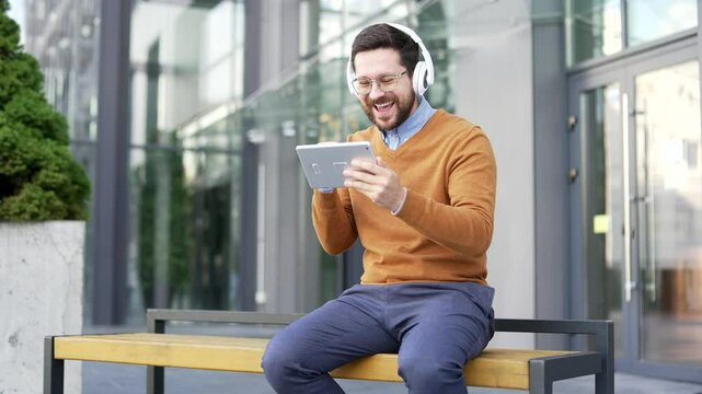 Excited businessman in headphones watching sports match or competition using tablet sitting on a bench on street near office building. Happy man cheering for bids at auction, celebrating success