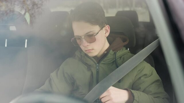 boy seen through windshield pulling seatbelt over shoulder inside parked car during winter, wearing green jacket and sunglasses, natural daylight and faint tree reflection on glass
