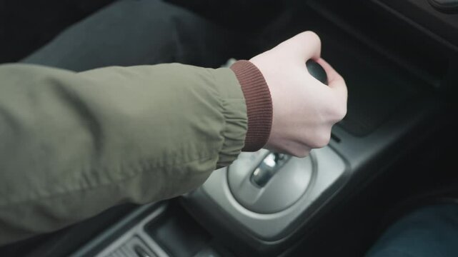 Close up top down view of driver pushing gear shift lever into park position in car center console showing hand grip on leather knob and metallic base under soft daylight in modern interior