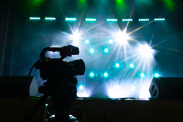 A dark silhouette of a professional video camera positioned in front of a bright stage illuminated by intense white and blue lights, surrounded by haze and visual depth.