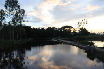 Obraz premium Calm Pond Reflecting Sky with Clouds, Trees, and Water Channel in Bangladesh Landscape
