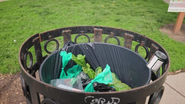 Close up of top of public outdoor trash-bin and dumpster can overfilled with compost and garbage with flies, bugs, stinky smells, and various trashy plastics and litter filling metal bucket at park