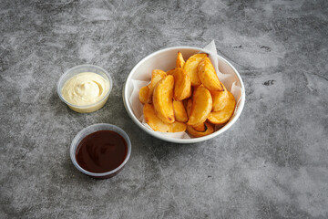 A bowl of fries with a dipping sauce and a container of ketchup. The fries are cut into wedges