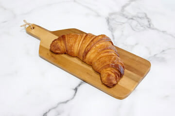 A croissant is sitting on a wooden cutting board. The croissant is brown and has a golden crust