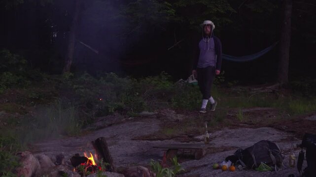 A man wearing a sweater and Tilly hat walks towards a campfire with a book at dusk. Hammocks are hung on trees in the background of this forested area.