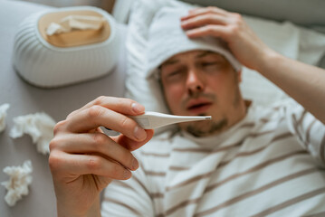 Sick man suffering from cold or flu, lying in bed with wet towel on his forehead, checking his body temperature with thermometer, tissues box and used tissues on the bed