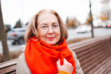 Senior mature smiling women 55-60 year old wearing vision glasses and red scarf with winter coat over city background. Wellness lifestyle. Looking at camera.