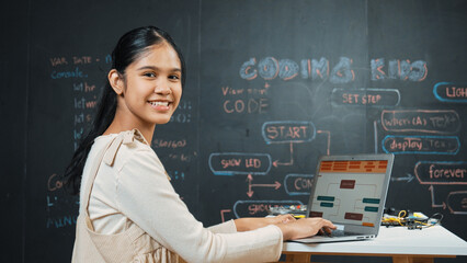 Caucasian girl working on laptop while smiling to camera at blackboard with engineering code and...