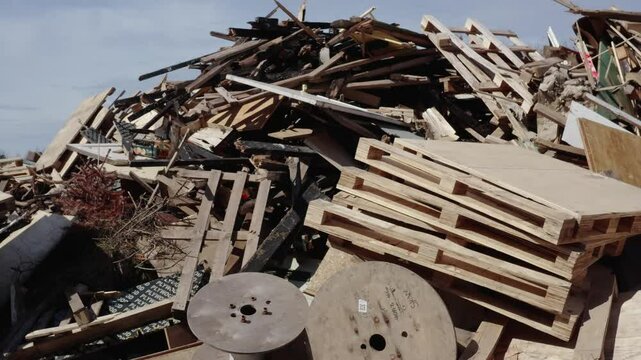 A large heap of discarded wooden pallets, construction timber, plywood, and cable reels piled in an industrial wood waste area under daylight.
