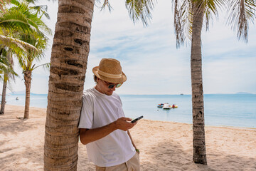 Smiling man using smartphone on tropical beach under shade of palm trees. summer holidays and people concept - portrait of happy smiling young man in sunglasses on the beach. Enjoying beach day