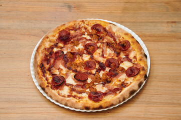 A pizza with pepperoni and ham on it. The pizza is on a white plate. The plate is on a wooden table