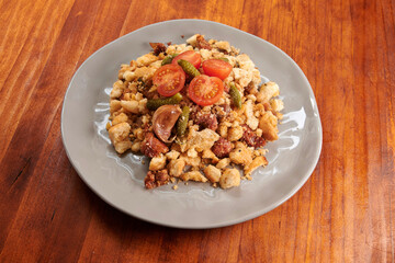 A plate of food with a tomato on top. The plate is on a wooden table. The food is a mix of meat and vegetables