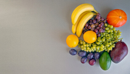 A colorful assortment of fruits with pumpkin on gray background. A vibrant display of fresh fruits, including bananas, grapes, oranges, plums, avocado, and a small pumpkin, arranged on a gray surface.